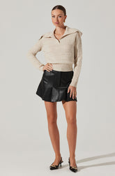 A woman is wearing a cream-colored, chunky knit sweater with a wide collar and long sleeves. She pairs it with a high-waisted black leather A-line mini skirt featuring vertical seam detailing and a slight flare. She completes the outfit with black pointed-toe high heels adorned with gold accents on the toes.